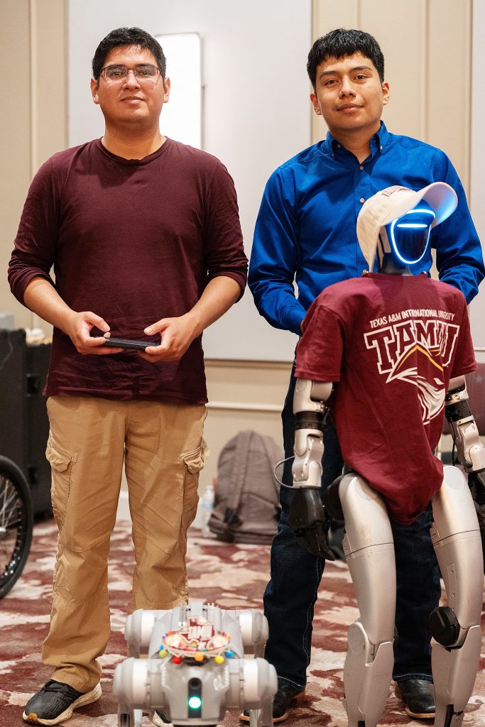 Two young men stand next to robots.