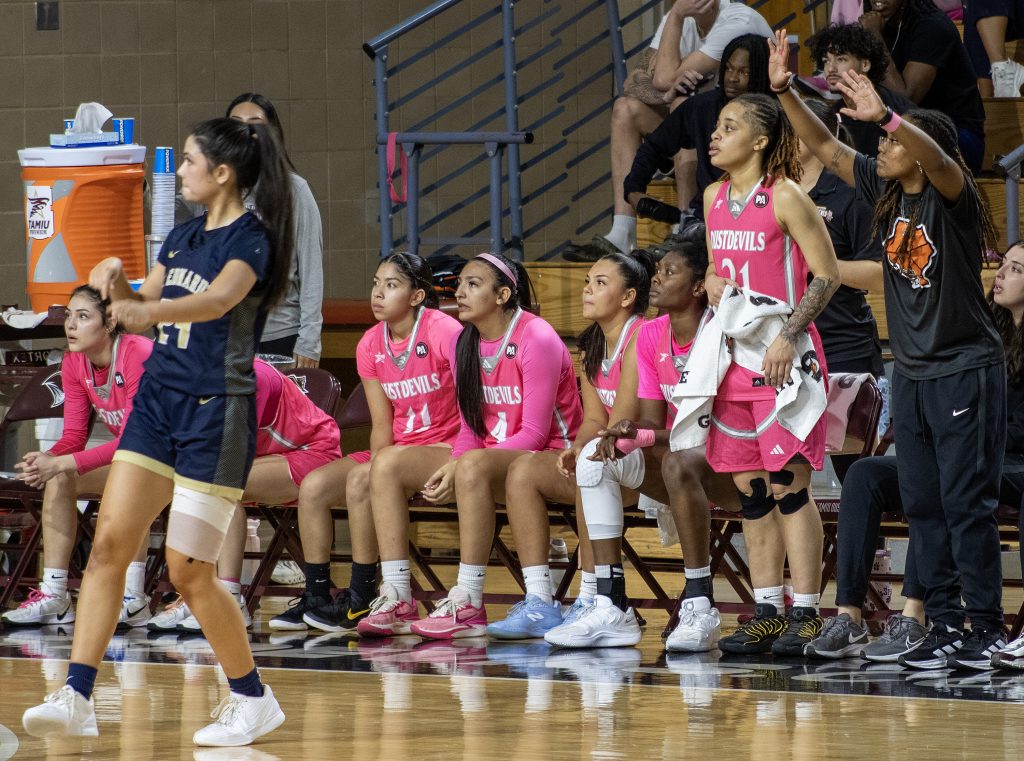 A group of women's basketball players wear pink jerseys as they sit on the bench and watch the game.