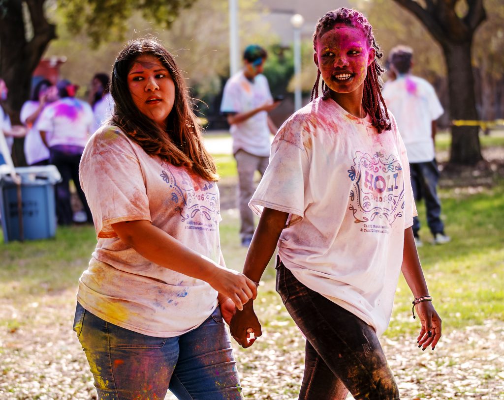 Two young women walk hand-in-hand, wearing white T-shirts covered in various colors of powder.