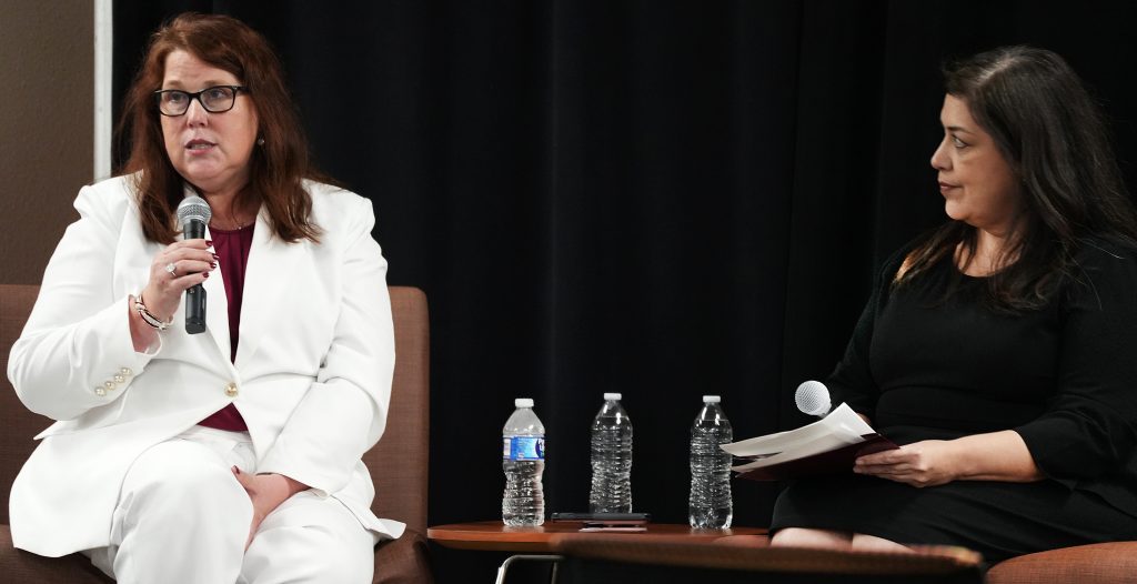 Two people sit during a forum. The one on the left is wearing a white suit and holding a microphone. The one on the right is dressed in black and listening to the other.